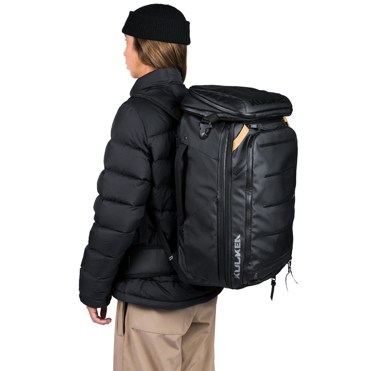 A person wearing a black puffer jacket, black beanie, and tan pants is shown from the back, carrying a large black Kulkaren backpack on their shoulders against a plain white background.
