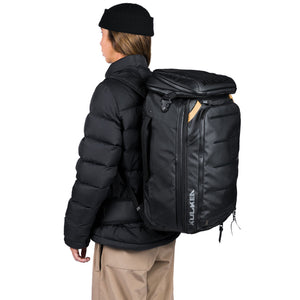 A person wearing a black puffer jacket, black beanie, and tan pants is shown from the back, carrying a large black Kulkaren backpack on their shoulders against a plain white background.