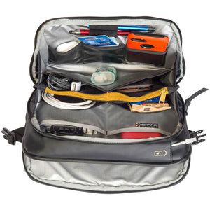 An open gray and black backpack shown from above, filled with various travel accessories including pens, a blue card, an orange case, a white cable, and a yellow zipper. The brand name 'KULLACK' is visible on a small tag inside the bag.