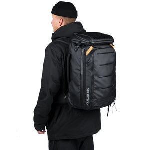A man wearing a black beanie and black jacket, shown from the back while wearing a black Aluxen backpack with tan accents.