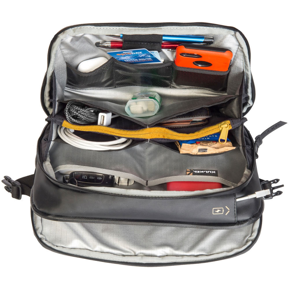 An open gray and black backpack shown from above, filled with various travel accessories including pens, a blue card, an orange case, a white cable, and a yellow zipper. The brand name 'KULLACK' is visible on a small tag inside the bag.