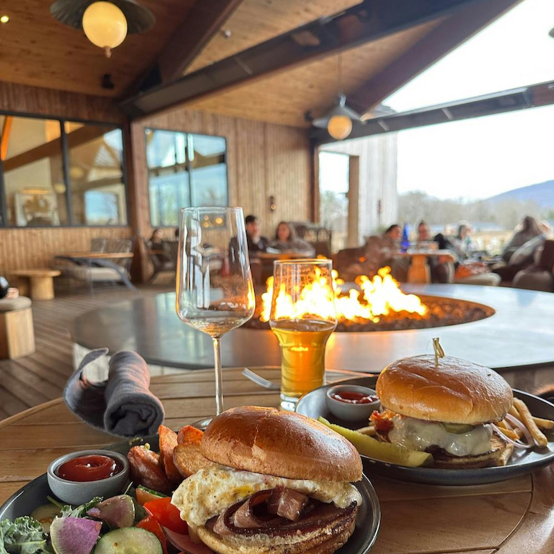 Restaurant table with sandwiches and drinks next to a fire with mountain view